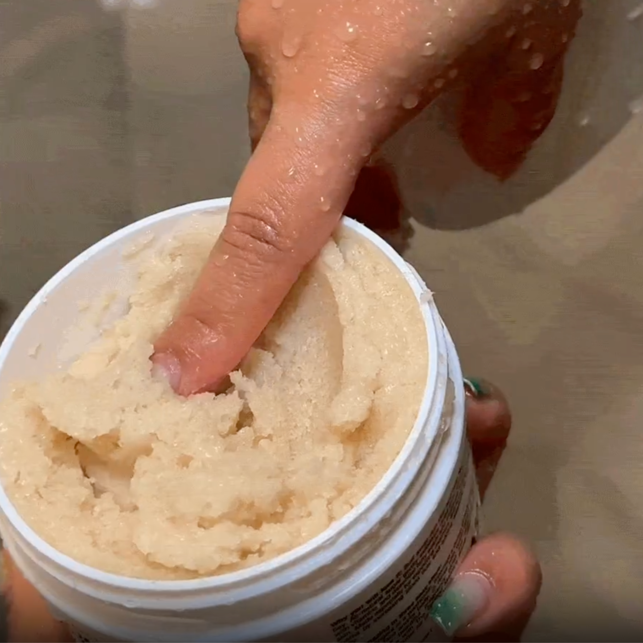 Hand holding a container of sugar scrub with a finger pressing into it, against a neutral background.